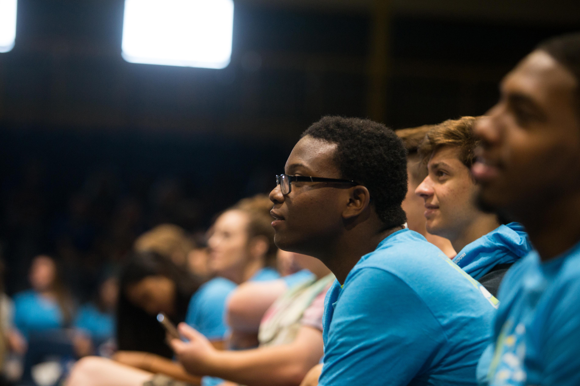 Black male student in glasses leans forward intently in row of students in blue shirts
