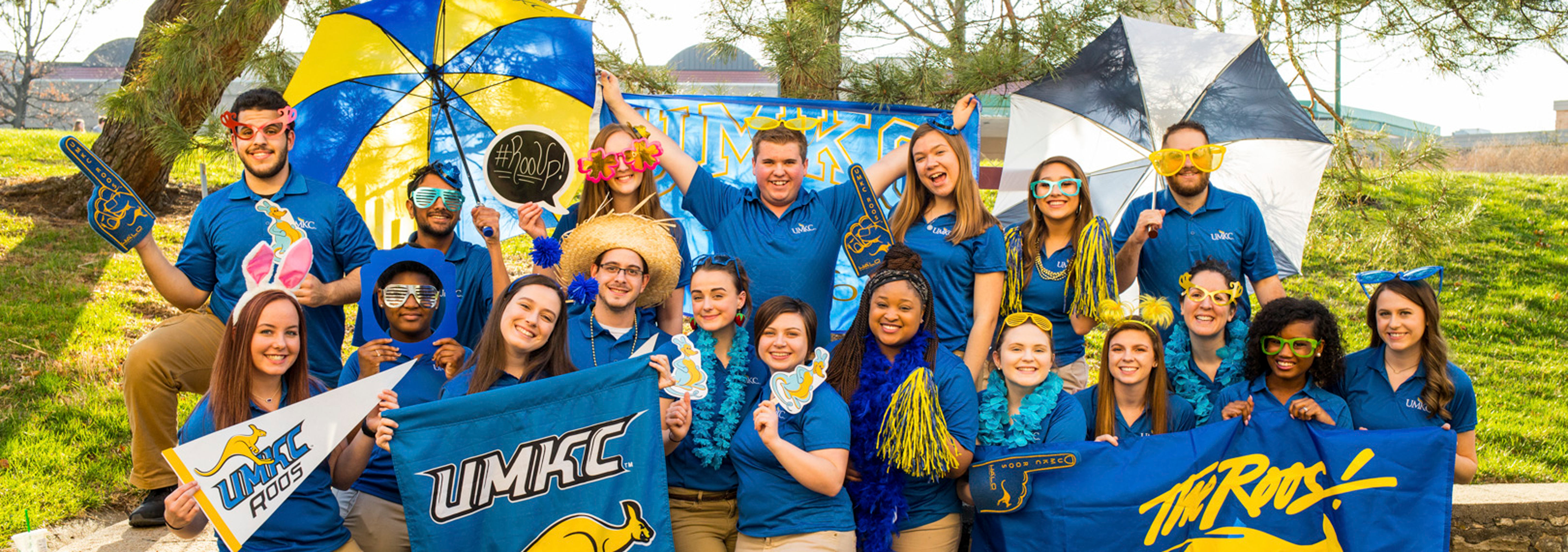 large group of diverse students with hats, big sunglasses and UMKC gear pose with UKMC flags
