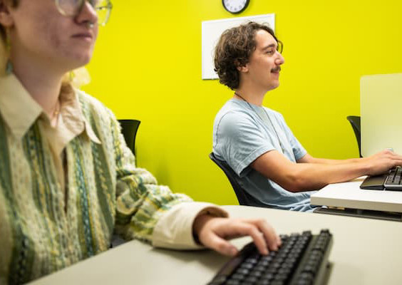 Two people are sitting at computer chairs. One has a mustache and short curly brown hair and is wearing a light blue shirt. The other is wearing glasses and a collared striped shirt.