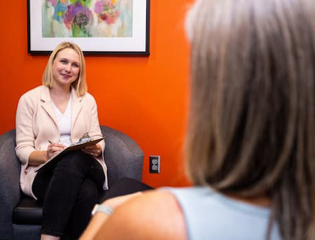 Person with blond hair is sitting in chair with clipboard talking to someone mostly out of frame who is also sitting.