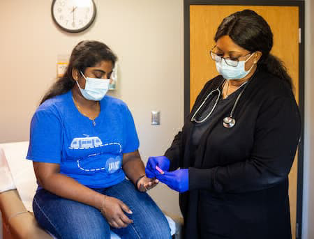 Roo Wellness staff member wearing scrubs and mask is pricking the finger of a patient who is also wearing a mask.