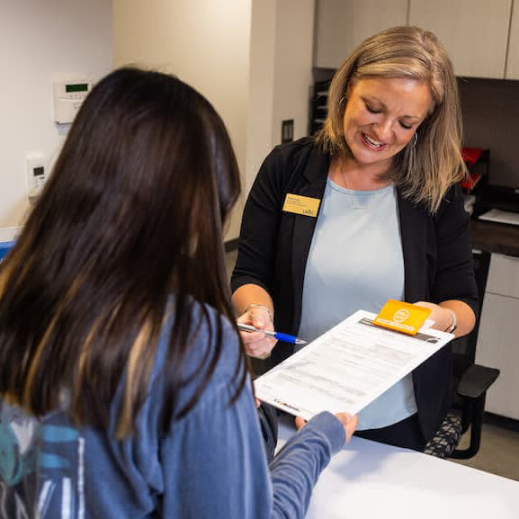 Person with blonde hair wearing black cardigan with nametag is helping person at desk and holding clipboard and pen