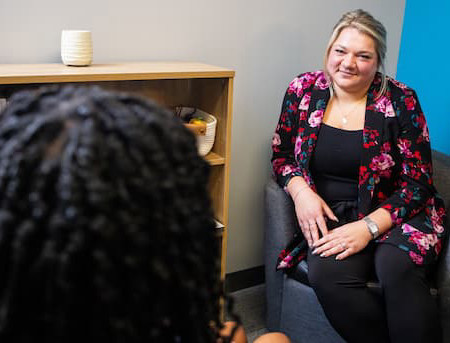 Person with blonde hair pulled back is sitting in chair talking with someone whose face is out of frame. The person with blonde hair is wearing black pants, black shirt and flower cardigan