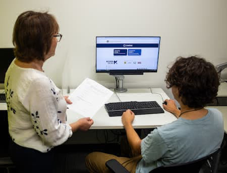 Staff member holding piece of paper sitting next to person at computer. 